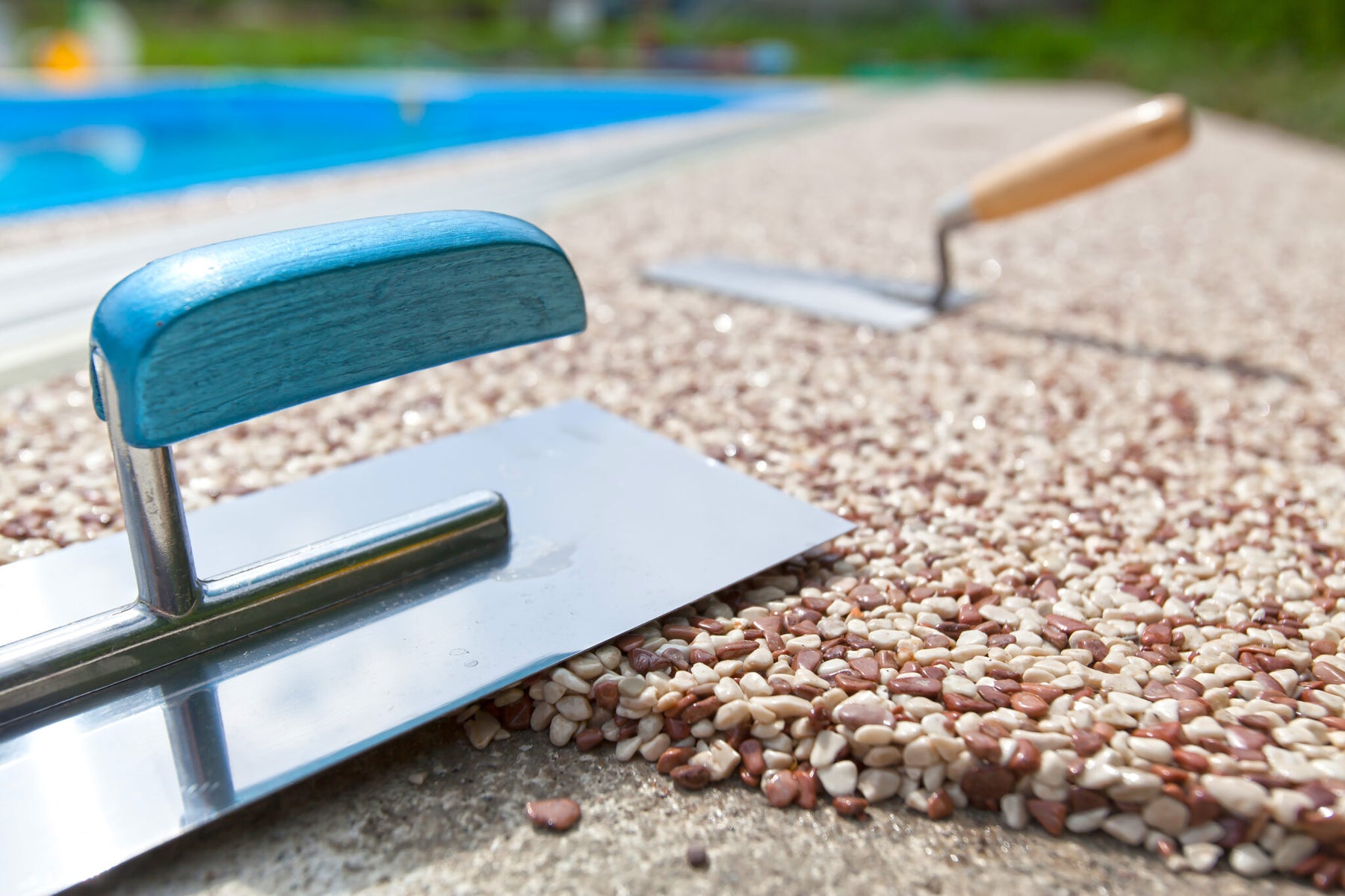 Colored exposed concrete aggregate being installed near a pool. There is a concrete float sitting on the aggregate. In the background, a concrete trowel is pressed against the exposed aggregate