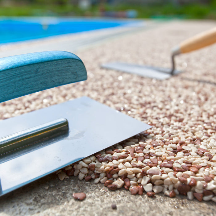 Colored exposed concrete aggregate being installed near a pool. There is a concrete float sitting on the aggregate. In the background, a concrete trowel is pressed against the exposed aggregate